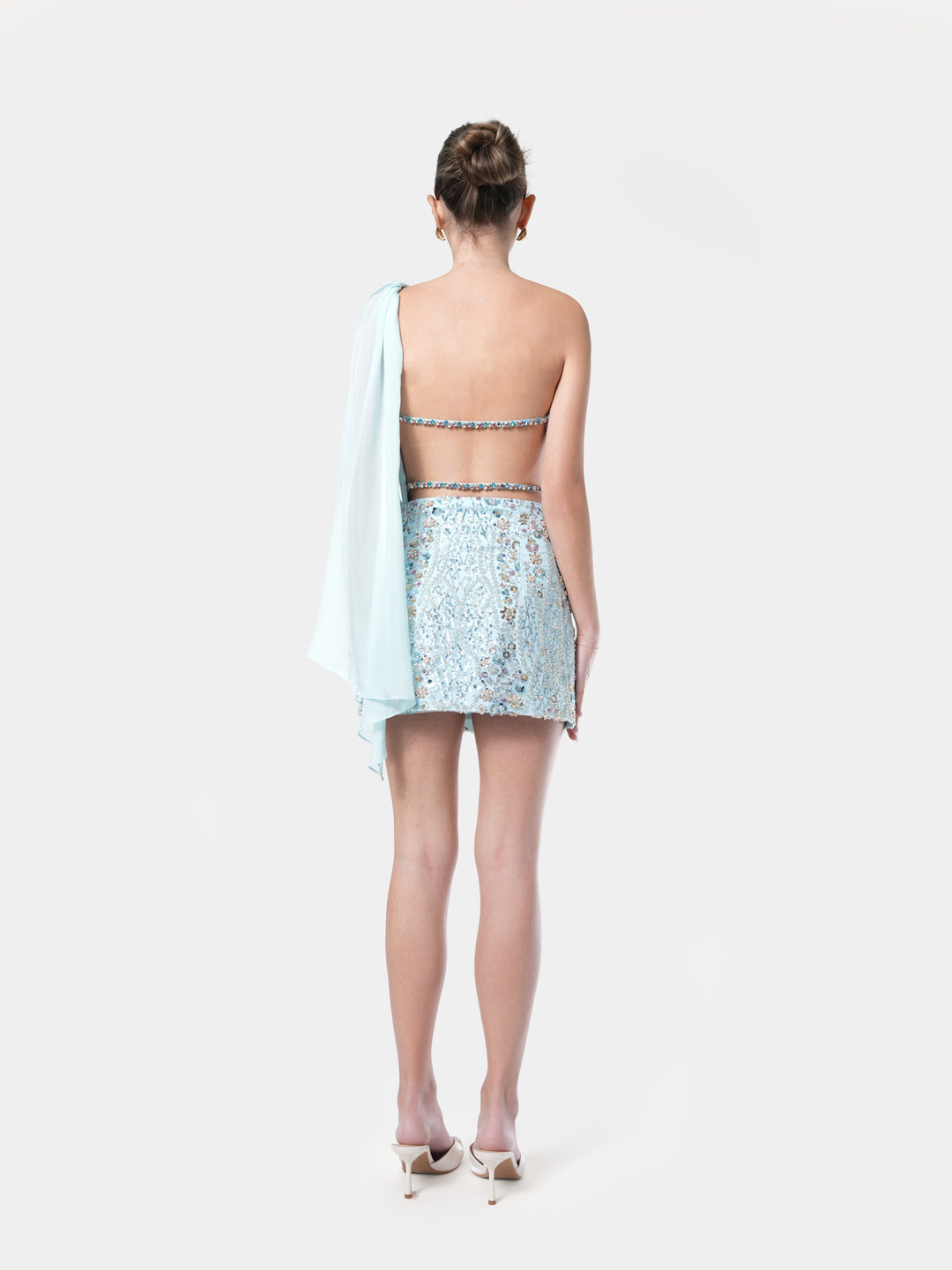 Woman wearing a light blue sequin top and skirt with a white shawl on a white background. NHÏJE aqua off shoulder top back view with crystals straps, premium craftsmanship, timeless elegance