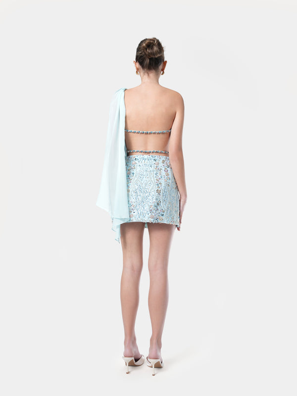Woman wearing a light blue sequin top and skirt with a white shawl on a white background. NHÏJE aqua off shoulder top back view with crystals straps, premium craftsmanship, timeless elegance